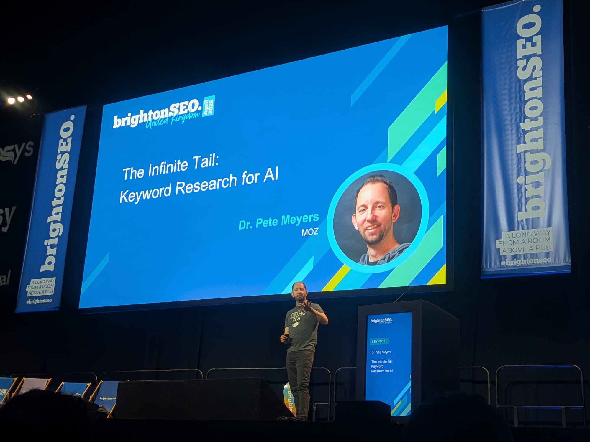 MozのDr. Pete MeyersがBrightonSEO Brighton 2026の基調講演「The Infinite Tail: Keyword Research for AI」のステージ上に登壇。Tシャツに「I'm here because I was told there would be tea(紅茶があると聞いたから来ただけだよ)」とティーポットのアイコン入り